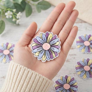 May include: A flower-shaped patch featuring alternating petals in shades of light purple, lavender, and pale yellow. The center of the flower is a textured, circular peach-colored disc. The entire design is outlined in a clean, white border.