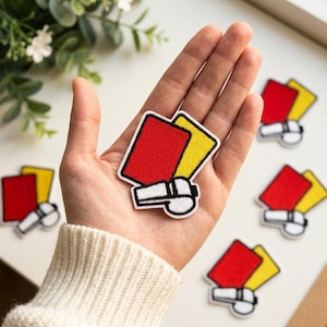 May include: An embroidered patch featuring a bright red rectangular card, a yellow rectangular card, and a white whistle with black outlines. The patch has a white border and is designed with a flat, stitched texture representing sports referee gear.