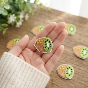 May include: A small, semi-circular embroidered patch featuring a kiwi fruit design. The patch displays a brown textured outer edge, a bright green inner section with black embroidered seeds, and a white border. Several identical patches are visible.