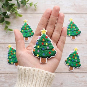 May include: A green embroidered Christmas tree patch featuring a yellow star at the top and colorful circular ornaments throughout the branches. The tree has a brown trunk and is outlined in white, with several identical patches visible nearby.