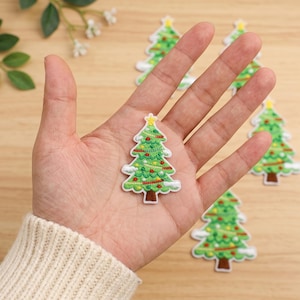 May include: A green embroidered Christmas tree patch featuring a yellow star topper, colorful ornaments, and white snow accents. The patch has a brown trunk and a white border. Several identical patches are visible in the background.