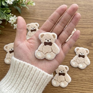 Peut inclure: Une collection de petits écussons en forme d'ours en peluche beige et duveteux avec des nœuds papillon brodés marron foncé. Chaque ours présente une surface texturée, des oreilles arrondies et un visage brodé simple.