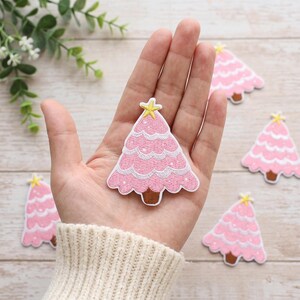 May include: An embroidered patch shaped like a Christmas tree in shades of pink with white scalloped trim, a brown trunk, and a yellow star at the top. The patch features a textured surface with visible stitching and is displayed in a palm.