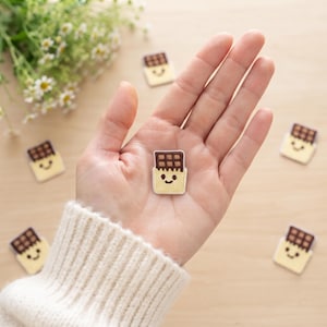 May include: A small, rectangular embroidered patch featuring a brown chocolate bar design with a light yellow wrapper. The patch has a stitched smiling face with two black eyes and a curved mouth. Several identical patches are scattered nearby.