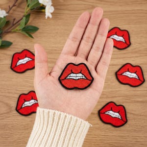 May include: A collection of embroidered patches shaped like red lips with white teeth and black outlines. Each patch features a vibrant red color for the lips, white thread detailing for the teeth, and a bold black border surrounding the entire shape.