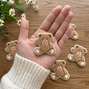 Könnte beinhalten: Ein brauner Teddybär-Aufnäher mit strukturierter, flauschiger Oberfläche, einer kleinen rosa Fliege um den Hals und weißen Akzenten an Ohren und Pfoten. Der Aufnäher zeigt einen sitzenden Bären, der winkend die Arme hebt.
