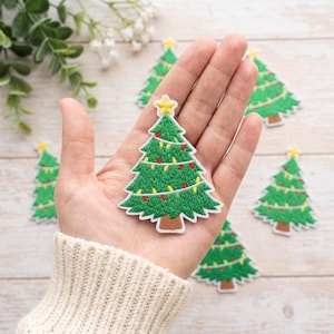 Puede incluir: Un parche bordado de árbol de Navidad verde con una estrella amarilla en la parte superior y luces de cadena coloridas en rojo y amarillo. El parche tiene un tronco marrón y un borde blanco, mostrando costuras detalladas en todo el diseño.