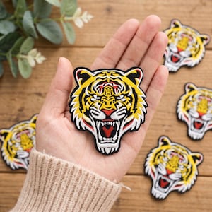 May include: An embroidered patch featuring a snarling tiger head with yellow, black, and white fur, a red nose, and an open mouth showing white teeth. The patch has a black border and is shaped like the tiger's head with pointed ears and whiskers.