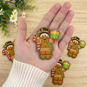 May include: An embroidered gingerbread person patch featuring a brown body, a black top hat, a red and white candy cane, a green and red striped scarf, and a green gift box with a white ribbon. The patch has decorative white icing details.