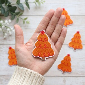 May include: An embroidered patch shaped like a stylized Christmas tree in shades of orange with a red star at the top and a brown trunk. The patch features a white border and decorative red swirl stitching. Several identical patches are visible.