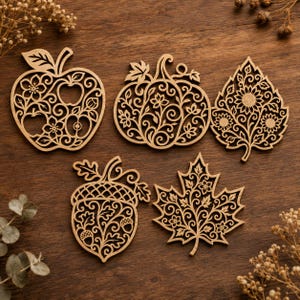 May include: Five laser-cut wooden ornaments featuring intricate filigree patterns. The shapes include an apple, a pumpkin, a sunflower-filled leaf, an acorn, and a maple leaf, all displaying detailed scrollwork and floral motifs in light brown.