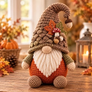 May include: A crochet gnome figurine featuring a tall brown textured hat adorned with a fabric orange maple leaf, a green leaf, a small red and white mushroom, and a tiny acorn. The gnome has a cream-colored yarn beard, a round nose, and an orange body.