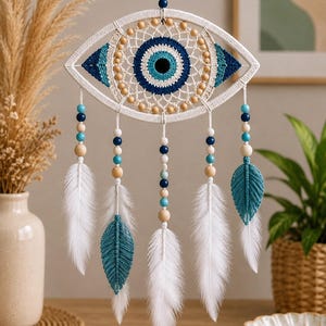 May include: A wall hanging featuring a central eye-shaped macrame frame with a blue and white iris design. The piece includes wooden and blue beads, white feathers, and two teal macrame leaf accents hanging from the bottom of the eye structure.