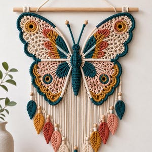 Puede incluir: Un tapiz de macramé con forma de mariposa, elaborado con cuerda anudada en tonos verde azulado, amarillo mostaza y crema. El diseño incluye patrones detallados en las alas, dos antenas con cuentas de madera y flecos de borlas en forma de hoja.