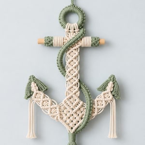 May include: A wall hanging in the shape of an anchor, crafted from cream and sage green macrame cord. The design features intricate knotted patterns, a central wooden dowel, and a decorative rope element that wraps around the anchor structure.