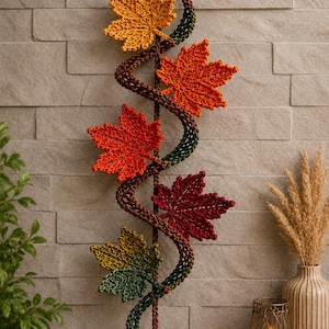 Op de afbeelding: Een verticale wandhanger met een centrale bruine gehaakte rank die naar beneden spiraalt, versierd met vijf gebreide esdoornbladeren in tinten geel, oranje, rood en groen, afgewerkt met een kleine bruine en gele gehaakte eikel aan de onderkant.