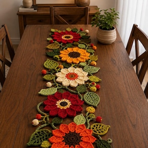 May include: A long, decorative table runner featuring a series of five large, crocheted flowers in shades of red, orange, yellow, and cream. The floral arrangement is accented with green crocheted leaves and small, round berry-like clusters.