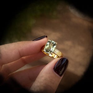 Könnte beinhalten: Ein Goldring mit einem großen, rechteckigen, hellgrünen Edelstein im Facettenschliff. Die Ringschiene ist aus poliertem Gold gefertigt und weist eine Reihe von vertikalen, parallelen Rillen oder Stegen auf, die den gesamten Ring umschließen.