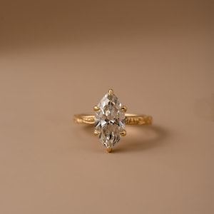 Puede incluir: Un anillo de oro con una piedra preciosa transparente de corte marquesa sujeta por seis garras. La banda del anillo presenta un patrón grabado y texturizado en su superficie, lo que aporta un acabado detallado a esta pieza de joyería.