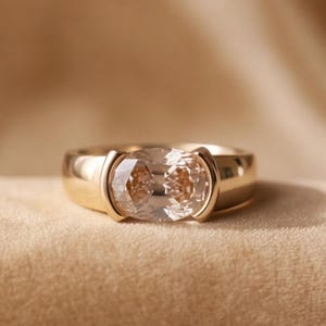 Puede incluir: Un anillo de oro con una piedra preciosa de color champán en forma ovalada horizontal, montada en un engaste de bisel. La banda es lisa y pulida, con el metal envolviendo los lados de la piedra central para asegurarla en su posición.