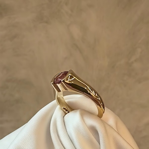 Può includere: Un anello in oro con una pietra preziosa rosa rotonda e sfaccettata incastonata in una fascia d'oro liscia e lucida. L'anello è ripreso da un'angolazione laterale, evidenziando la superficie metallica riflettente e la tonalità rosa vivace.