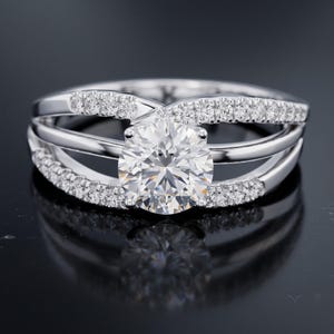 Könnte beinhalten: Ein silberfarbener Ring mit einem großen, runden, facettierten Mittelstein in einer mehrsträngigen Fassung. Die Ringschiene besteht aus drei einzelnen Strängen, wobei der obere und der untere Strang mit kleinen, klaren Steinen besetzt sind.