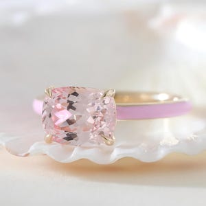 Peut inclure: Une bague en or ornée d'une grande pierre précieuse rose pâle taille coussin, fixée par quatre griffes. L'anneau de la bague est recouvert d'un émail rose clair lisse qui entoure le doigt, offrant une apparence nette et polie.