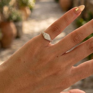 Può includere: Un anello color argento con una parte frontale a forma di marquise incastonata con molteplici piccole pietre chiare e scintillanti. La fascia è sottile e metallica, progettata per adattarsi al dito, mostrando una superficie dettagliata e testurizzata.