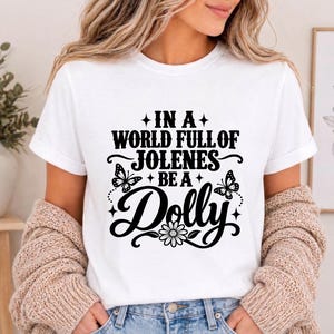 Puede incluir: Una camiseta blanca de manga corta con texto negro que dice IN A WORLD FULL OF JOLENES BE A Dolly. El diseño incluye dos mariposas, cuatro pequeñas estrellas de cuatro puntas y un gráfico de una margarita centrado bajo el texto.