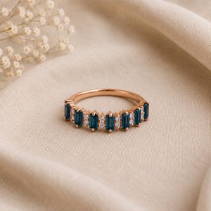 May include: A rose gold ring featuring a row of alternating dark blue baguette-cut stones and small round clear stones. The band has a polished metallic finish and is set with five rectangular blue gems separated by pairs of tiny white circular stones.
