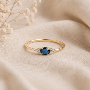 Puede incluir: Un anillo fino de oro con un zafiro azul central de talla ovalada, flanqueado por tres pequeños diamantes transparentes de talla redonda a cada lado, montados en un engaste de garras sobre una delicada banda de oro pulido.