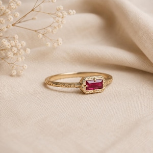 May include: A gold ring featuring a rectangular, baguette-cut deep red ruby gemstone set in a decorative, engraved metal bezel. The band of the ring displays a continuous, intricate scrollwork pattern etched into the surface of the gold metal.