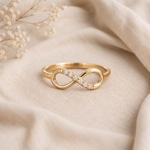 Op de afbeelding: Een gouden ring met een oneindigheidssymbool. De ringband is van gepolijst goud en een zijde van de oneindigheidslus is bezet met vijf kleine, heldere, rondgeslepen steentjes die schitteren. De ring heeft een gladde, doorlopende band.
