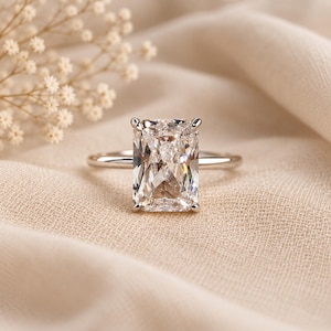 May include: A radiant-cut clear gemstone ring set in a polished silver-toned metal band. The rectangular stone features multiple facets that reflect light, held securely by four small prongs on a smooth, thin, circular band of metallic material.
