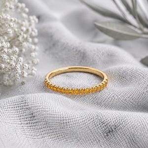 May include: A thin gold band ring featuring a continuous row of small, round, vibrant yellow gemstones set along the outer circumference. The metal has a polished finish, and the stones are uniform in size, creating a delicate, circular design.
