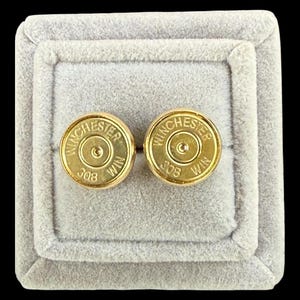 May include: A pair of circular gold-toned cufflinks crafted from the base of Winchester 308 WIN ammunition cartridges. Each piece features the embossed text Winchester and 308 WIN around the central primer, presented in a light gray velvet box.