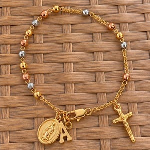 May include: A tri-colour beaded bracelet featuring alternating gold, rose gold, and silver-toned faceted beads. The bracelet includes a gold-toned lobster clasp, a dangling crucifix charm, a circular religious medal, and a letter A charm.