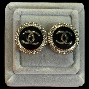 May include: A pair of round earrings featuring a black circular center with a gold-toned interlocking CC logo. The earrings are framed by a textured gold-toned rope-style border, creating a distinct contrast between the dark center and the metallic edge.