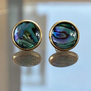 May include: A pair of round stud earrings featuring iridescent abalone shell inlays set within smooth gold-toned metal bezels. The natural shell displays swirling patterns of deep blue, green, and purple hues, creating a reflective surface.