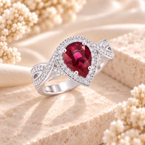 May include: A silver-toned ring featuring a pear-shaped deep red gemstone at the center, surrounded by a halo of small white stones. The band is split into two intertwining sections, both encrusted with small white stones, creating a twisted design.