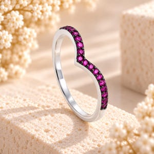 May include: A silver V-shaped ring featuring a row of small, round, vibrant pink stones set along the top curve. The band has a polished metallic finish, and the stones are held in place by dark, contrasting prongs, creating a distinct chevron design.