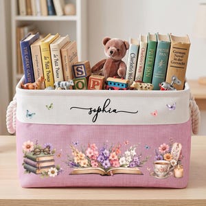 Peut inclure: Un bac de rangement rectangulaire en tissu rose avec des poignées en corde blanche, portant le prénom Sophia en écriture noire sur une bande supérieure blanche. L'avant présente des illustrations d'un livre ouvert avec des fleurs colorées.
