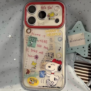 Puede incluir: Una funda transparente para smartphone con un marco de cámara rojo y varias pegatinas de Snoopy y Woodstock. El diseño incluye textos como "let's Go!", "Home is WHERE THE DOG", "SNOOPY DROPS" y "TAKE ME". La funda muestra personajes de dibujos.