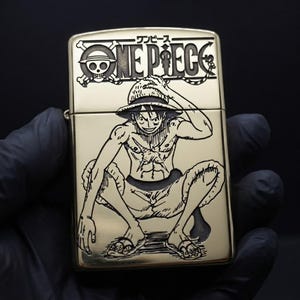 May include: A gold-toned metal lighter featuring a laser-engraved illustration of the character Monkey D. Luffy from the anime series One Piece. The top portion displays the One Piece logo with Japanese text, while the lower section shows Luffy.