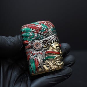 Puede incluir: Una funda de encendedor rectangular de metal con un diseño en relieve tridimensional de una figura con un tocado de plumas. El tocado está decorado con acentos de esmalte rojo y verde, mientras que el rostro y la base tienen un acabado dorado.