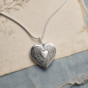 May include: A silver-coloured heart-shaped locket pendant suspended from a matching thin snake chain. The locket features an intricate embossed floral pattern surrounding a smooth, plain heart shape at its centre, creating a detailed metallic surface.