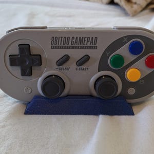 Puede incluir: Un mando de juego Bluetooth 8BitDo gris con una cruceta negra, dos joysticks analógicos grises y cuatro botones de acción de colores etiquetados como X, Y, A y B. El mando incluye botones Select y Start y descansa sobre un soporte azul.