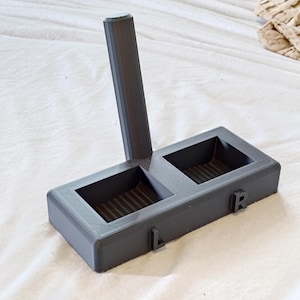 May include: A dark grey 3D printed stand featuring a vertical cylindrical post attached to a rectangular base with two square recessed compartments. The base has the letters L and R embossed on the front edge, indicating left and right sides.