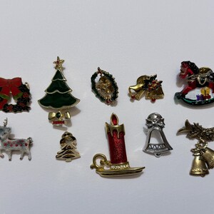 May include: A collection of ten Christmas-themed metal enamel pins featuring a red wreath, a green Christmas tree, a small wreath, gold bells, a rocking horse, a white reindeer, a gold tree, a red candle, a silver bell, and gold hanging bells.