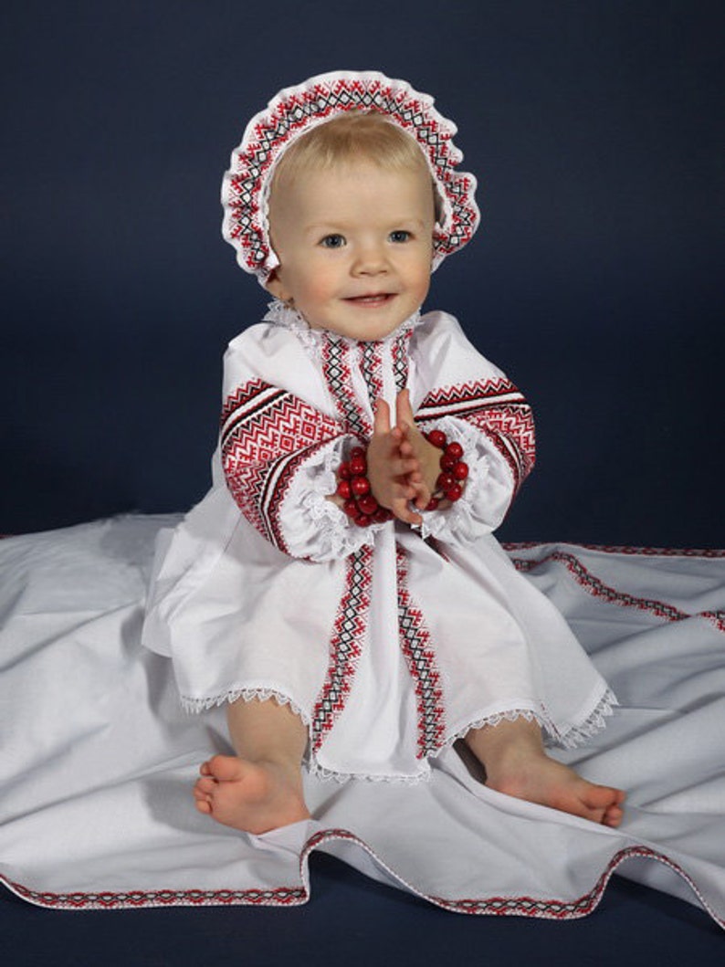 May include: A baby in a white dress and bonnet, both featuring red and black embroidery. The dress has a ruffled collar and long sleeves. The baby sits on a white cloth with embroidered edges, wearing a red beaded bracelet.